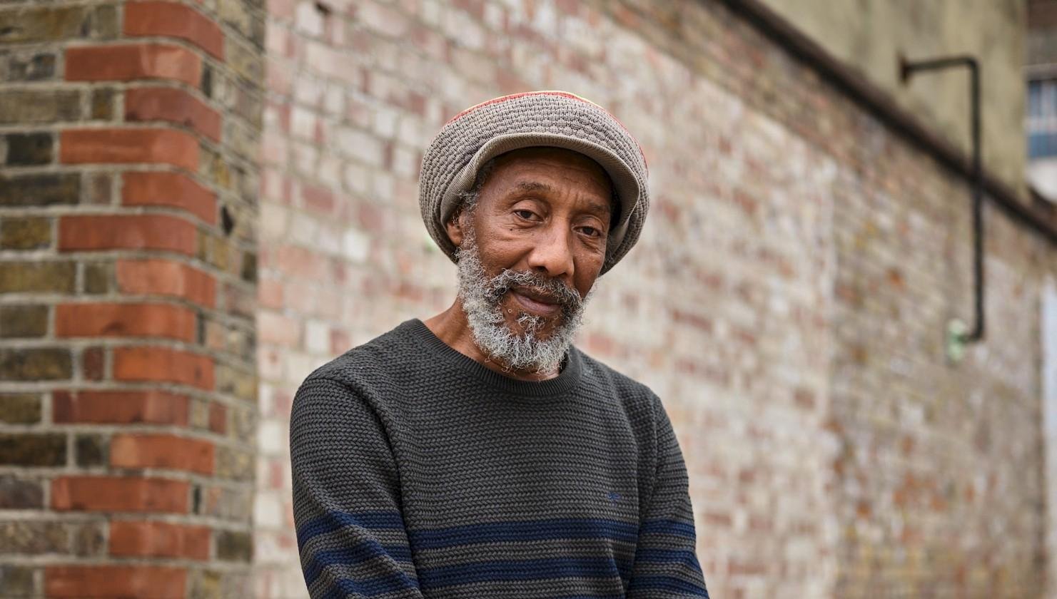Colour photograph of Jamaican reggae artist Locksley Gichie, wearing a striped jumper, jeans and a grey woollen hat. He is sitting on a stack of wooden pallets in front of a red brick wall.